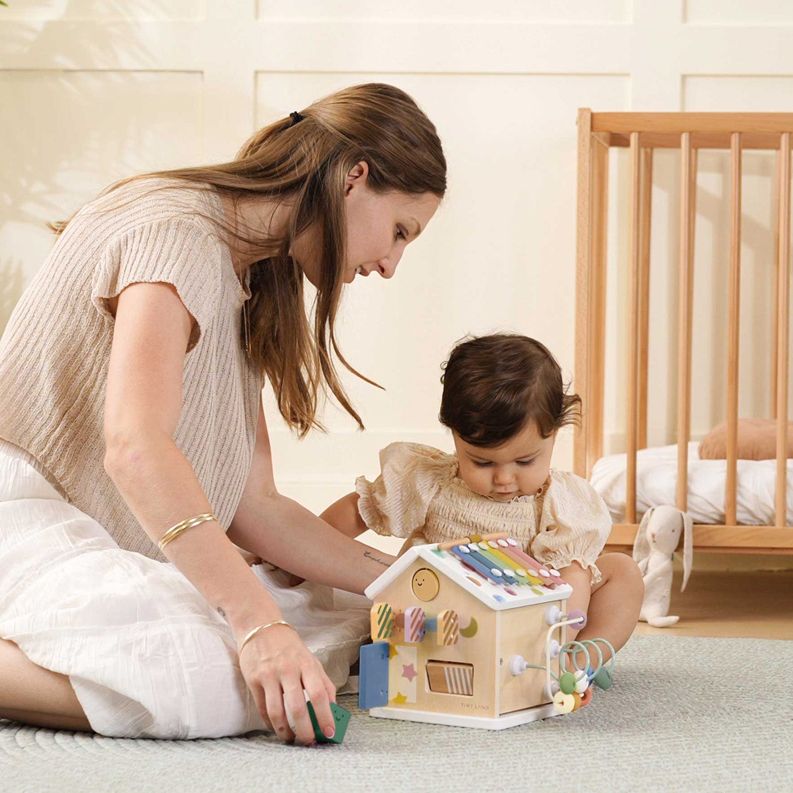 A woman and a young child sit on the floor, playing with the Tiny Land Cozy Learning Playset—Playmat + Activity Cube. A crib and stuffed animal are in the background, complementing this colorful developmental toy.
