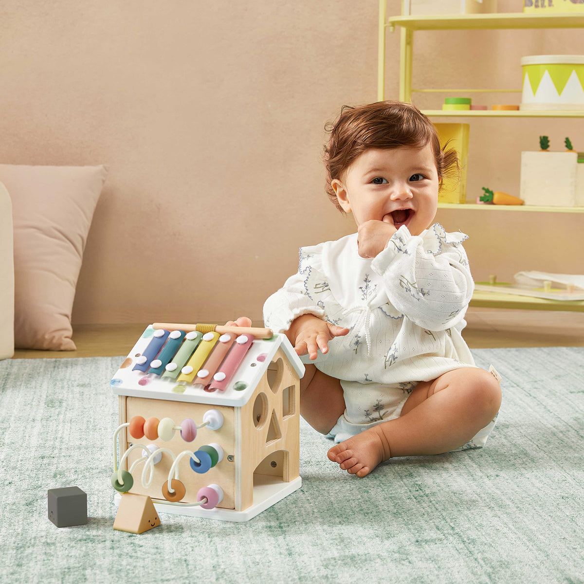 A smiling baby in a white outfit sits on the Cozy Learning Playset—Playmat + Activity Cube by Tiny Land, playing on the mat with the house-shaped activity cube. In the background, a yellow shelf holds various toys and decorations.