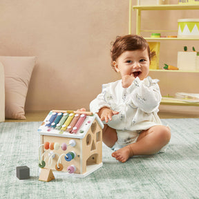 A smiling baby in a white outfit sits on the Cozy Learning Playset—Playmat + Activity Cube by Tiny Land, playing on the mat with the house-shaped activity cube. In the background, a yellow shelf holds various toys and decorations.