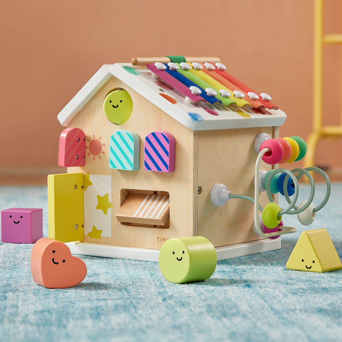 The Tiny Land Rainbow Baby Walker & Activity Cube Developmental Playset—a vibrant wooden house with a xylophone roof, shape sorter, bead maze, and smiley faces—boosts fine motor skills and is set on a blue carpet with a peach-colored background.