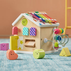 The Tiny Land Rainbow Baby Walker & Activity Cube Developmental Playset—a vibrant wooden house with a xylophone roof, shape sorter, bead maze, and smiley faces—boosts fine motor skills and is set on a blue carpet with a peach-colored background.