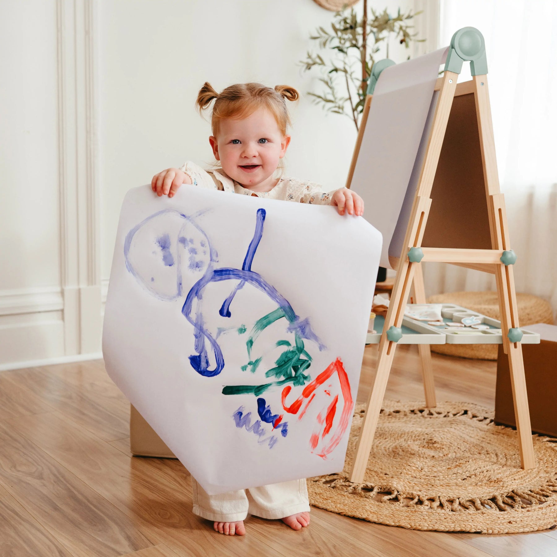 A toddler with pigtails smiles, holding a colorful painting beside the Tiny Land Nature-inspired Play & Art Set — Sage Easel + Forest Play House in a cozy, sunlit room with wooden floors.|green