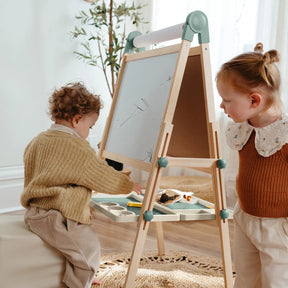 Two children create with the Tiny Land Nature-inspired Play & Art Set — Sage Easel + Forest Play House. One draws while the other observes, surrounded by art supplies and nature-themed decor for imaginative play.|green