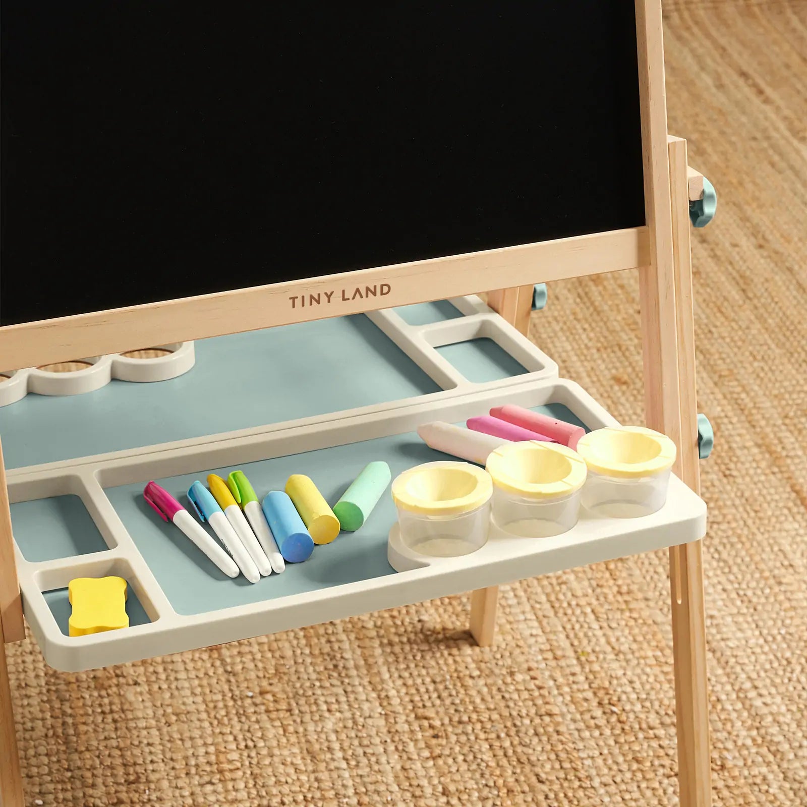 The Tiny Land Nature-inspired Play & Art Set — Sage Easel + Forest Play House stands on a woven rug, with a chalkboard, markers, colored chalk, yellow eraser, and three small containers neatly arranged on its tray.|green