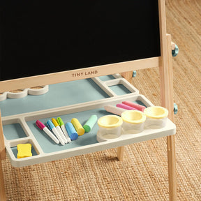 The Tiny Land Nature-inspired Play & Art Set — Sage Easel + Forest Play House stands on a woven rug, with a chalkboard, markers, colored chalk, yellow eraser, and three small containers neatly arranged on its tray.|green