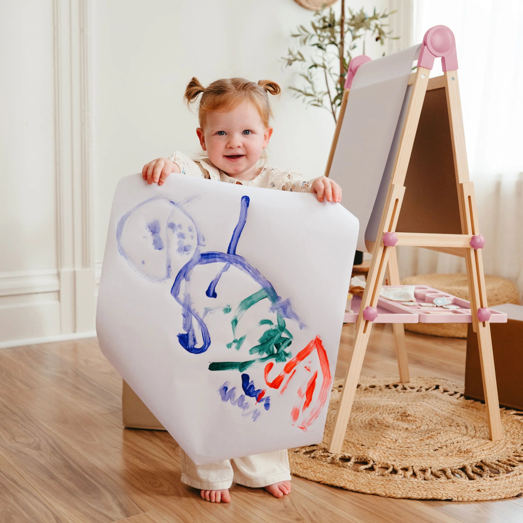 A young child smiles and displays colorful abstract art while standing next to the Tiny Land® Magnitales Easel for Kids - Pink by Tiny Land in a cozy, well-lit room.|pink