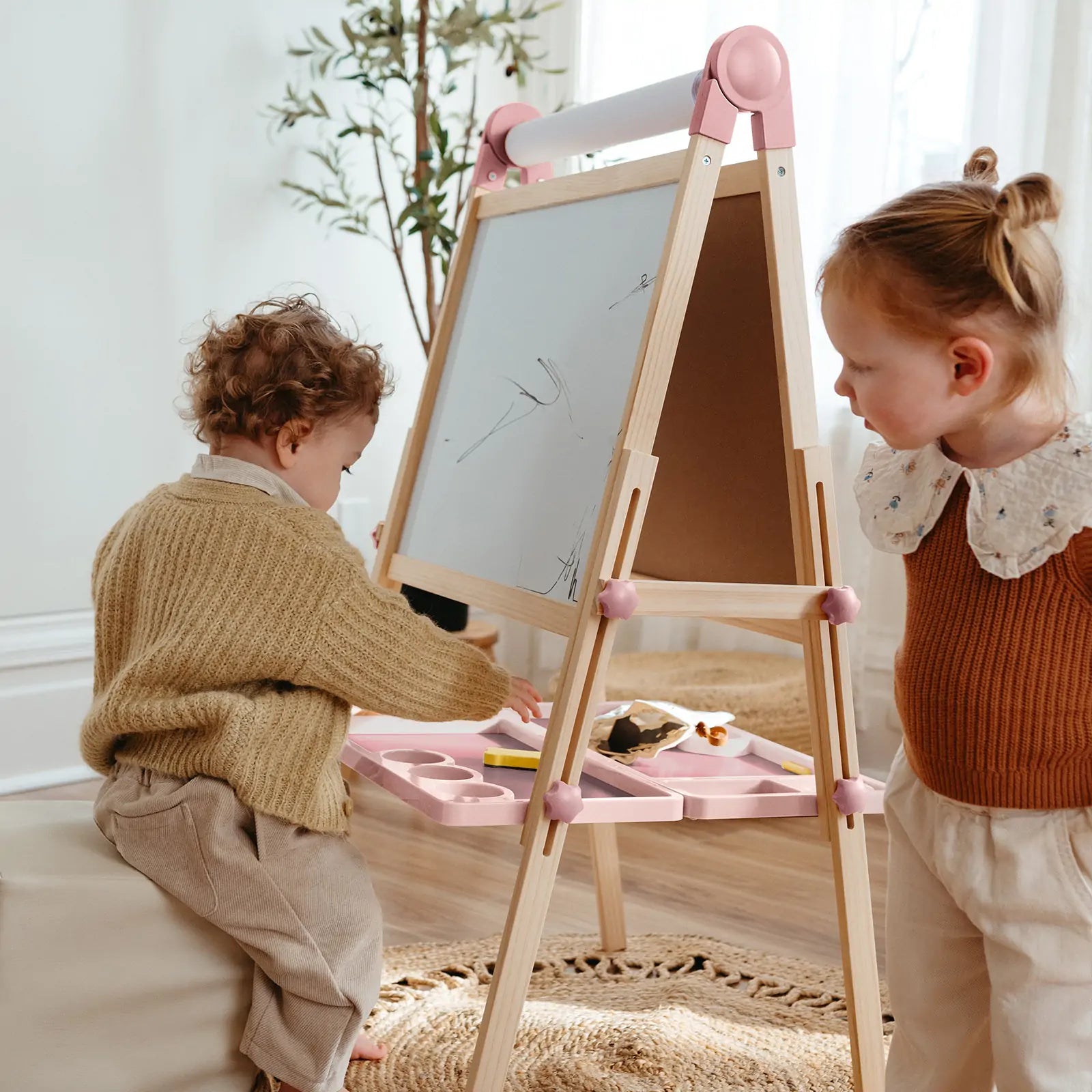 Two children draw and use art supplies together on the Tiny Land® Magnitales Easel for Kids - Pink, a height-adjustable whiteboard easel with tray, in a cozy room with sunlight, a rug, and a plant.|pink