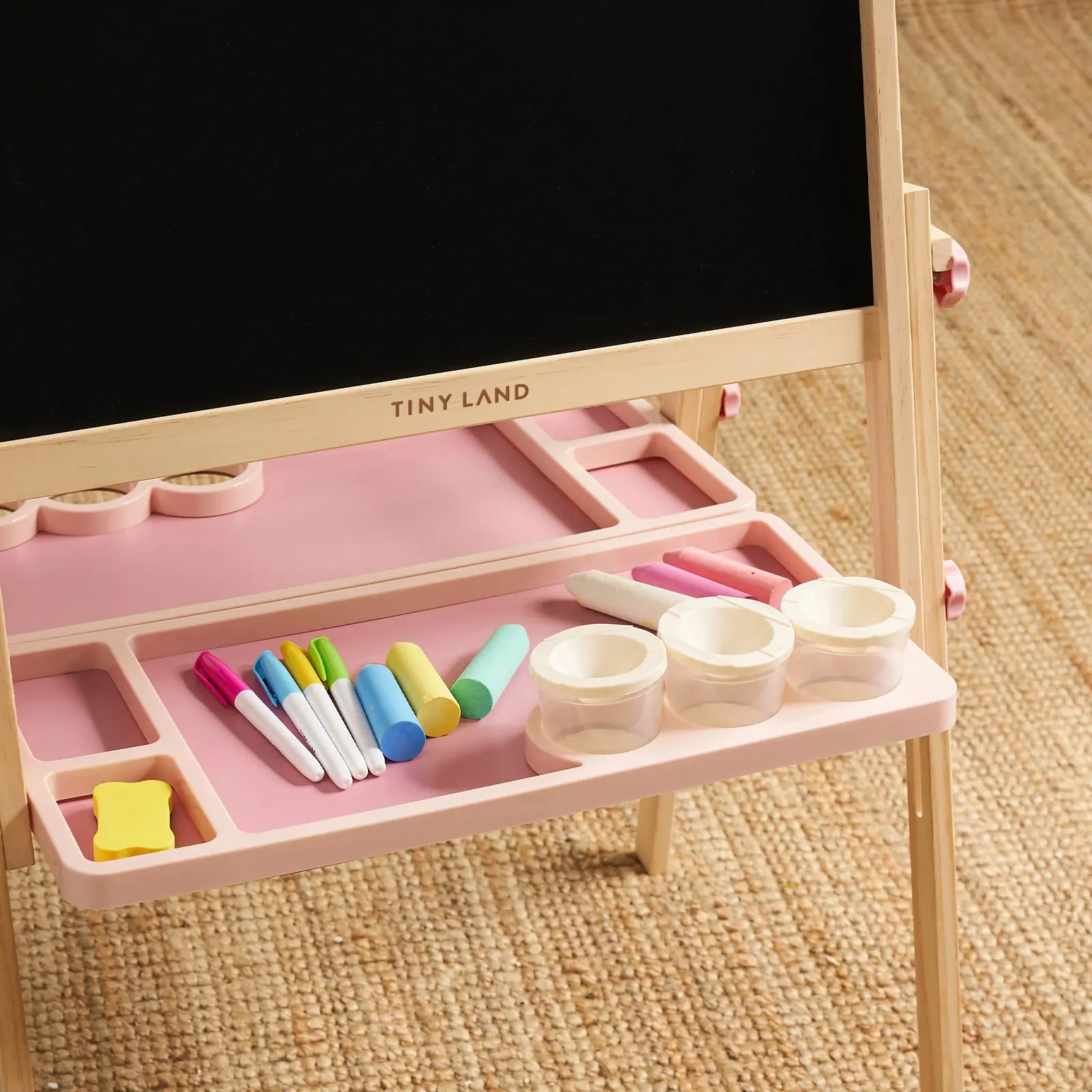 The Tiny Land® Magnitales Easel for Kids - Pink features a height adjustable blackboard, wooden legs, and tray to hold markers, chalk, a yellow eraser, and three clear cups. "Tiny Land" is printed on the wood. The easel sits on a woven carpet.|pink
