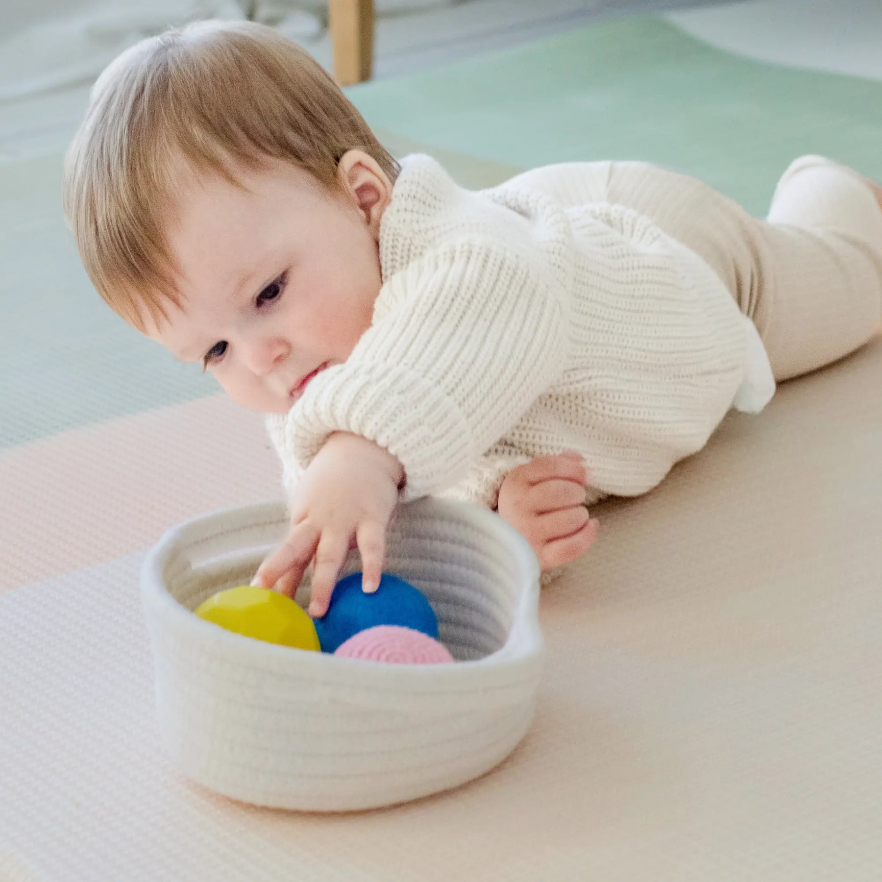 A baby in a cream sweater enjoys sensory exploration and fine motor play with the Tiny Land® Montessori Early Skills Baby Set (7–12 months) by Tiny Land, reaching for colorful balls on a soft mat from a white basket.