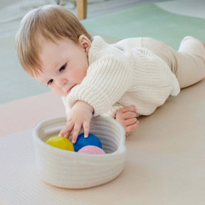 A baby in a cream sweater enjoys sensory exploration and fine motor play with the Tiny Land® Montessori Early Skills Baby Set (7–12 months) by Tiny Land, reaching for colorful balls on a soft mat from a white basket.
