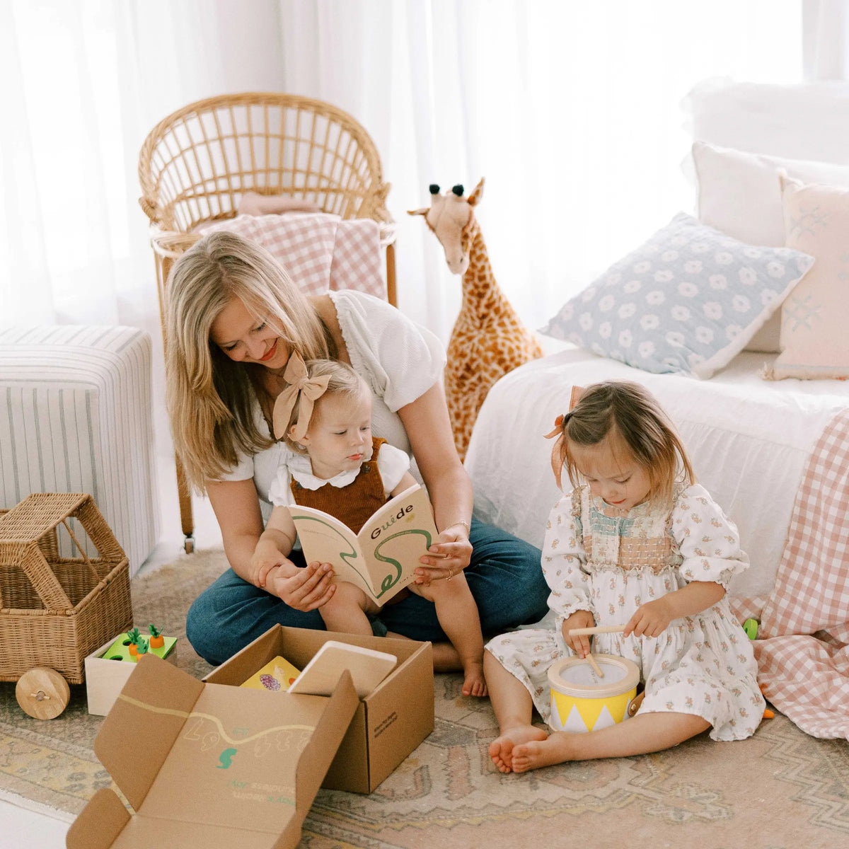 A woman sits on the floor with two young children in a sunlit room. One, about 13–15 months old, sits on her lap with a book, while the other plays a drum near Tiny Land® Montessori Toys for Toddlers and a large giraffe plush.