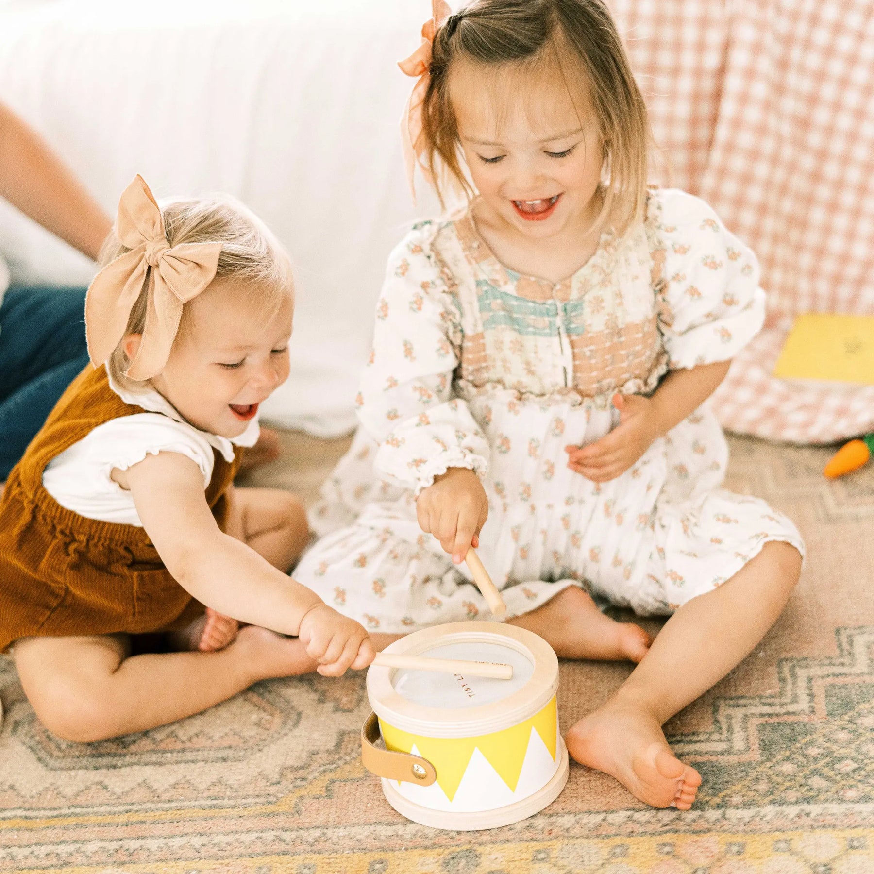 Two young children smile as they play with the Tiny Land® Montessori Toys for Toddlers (13–15 Months) drum, each holding a drumstick. One wears a white floral dress, the other a brown pinafore and bow headband. Brand: Tiny Land.