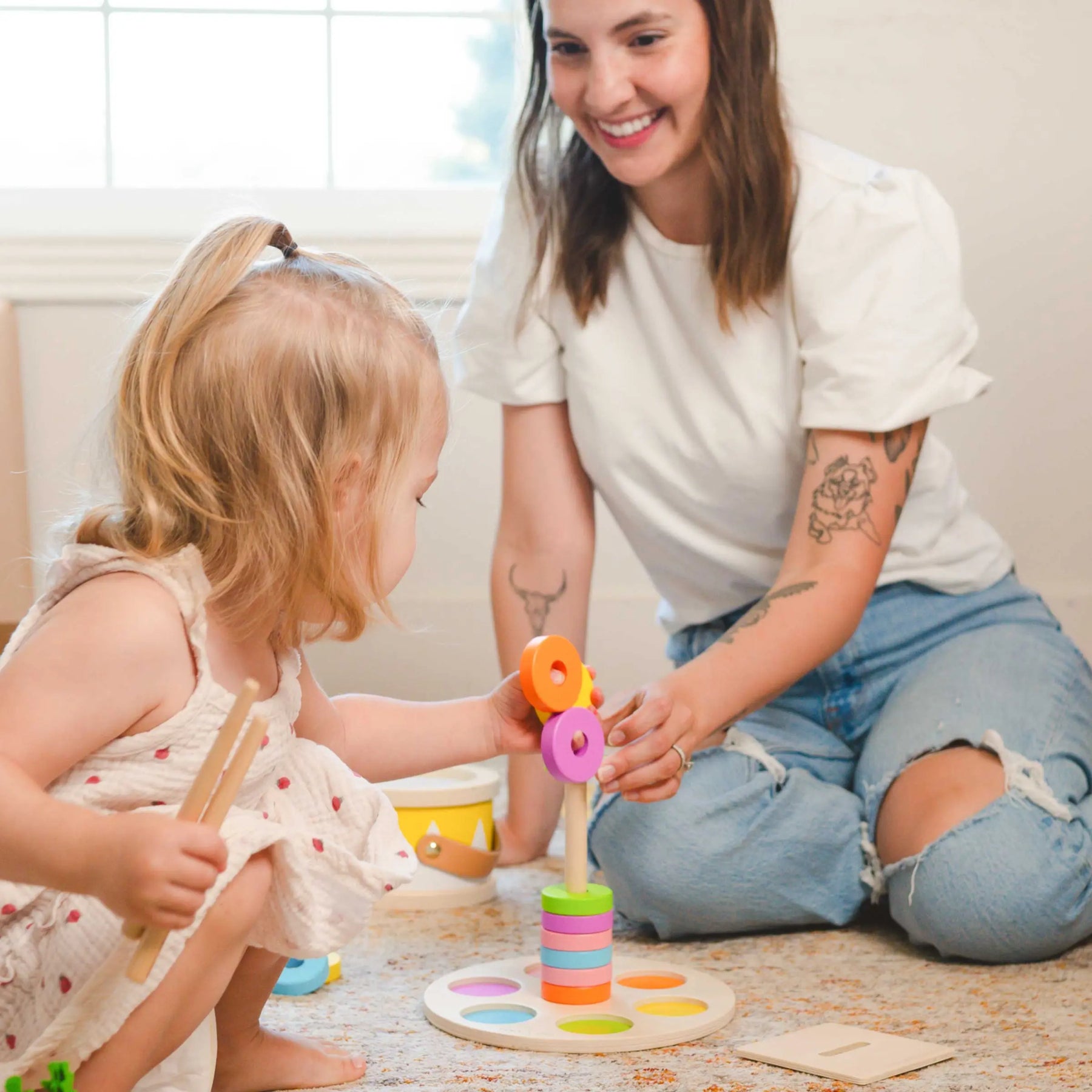 A young woman smiles as she and a toddler stack colorful rings from the Tiny Land® Montessori Advanced Skills Baby Set (13–24 months), encouraging fine motor skills together in a bright, cozy room.