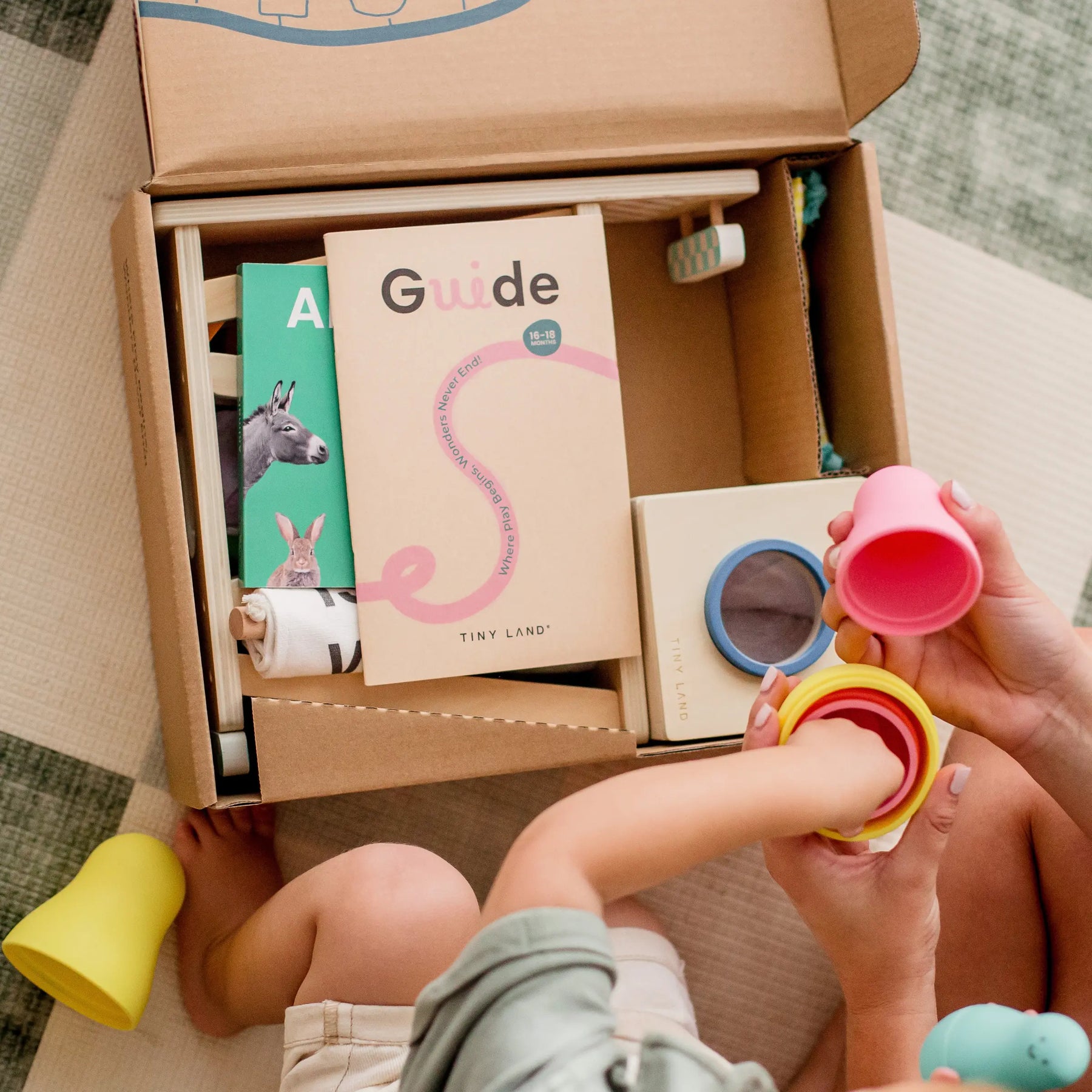A child and adult enjoy Tiny Land® Montessori Toys for Toddlers (16–18 Months) alongside an open box with books, a guide, and educational toys—perfect for curious minds. From Tiny Land.