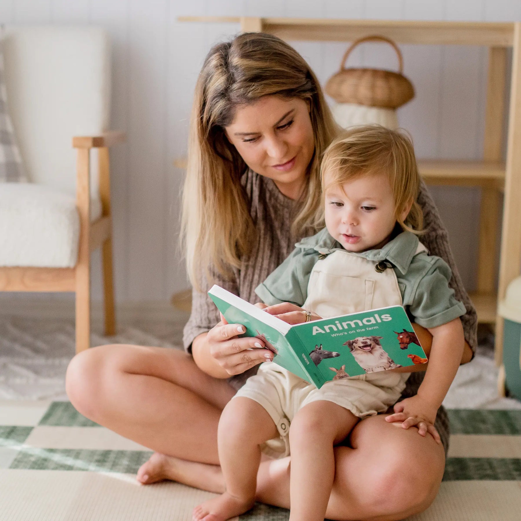 A woman sits on the floor with a young child on her lap, reading and exploring the Tiny Land® Montessori Advanced Skills Baby Set (13–24 months) by Tiny Land in a cozy, softly lit room to nurture sensory and motor development.