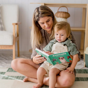 A woman sits on the floor with a young child on her lap, reading and exploring the Tiny Land® Montessori Advanced Skills Baby Set (13–24 months) by Tiny Land in a cozy, softly lit room to nurture sensory and motor development.