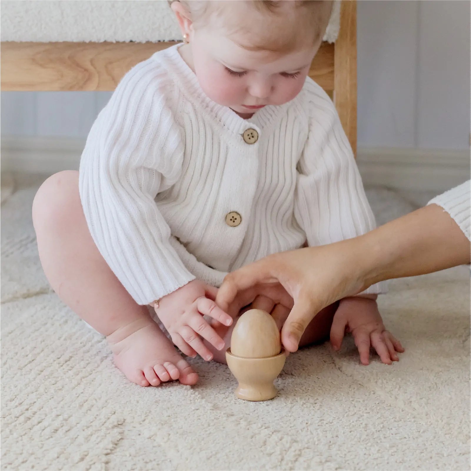 A baby in a white sweater sits on a cream carpet as an adult offers the Tiny Land® Montessori Toys for Infants (10–12 Months), sparking curiosity and discovery through Tiny Land’s baby development toys.