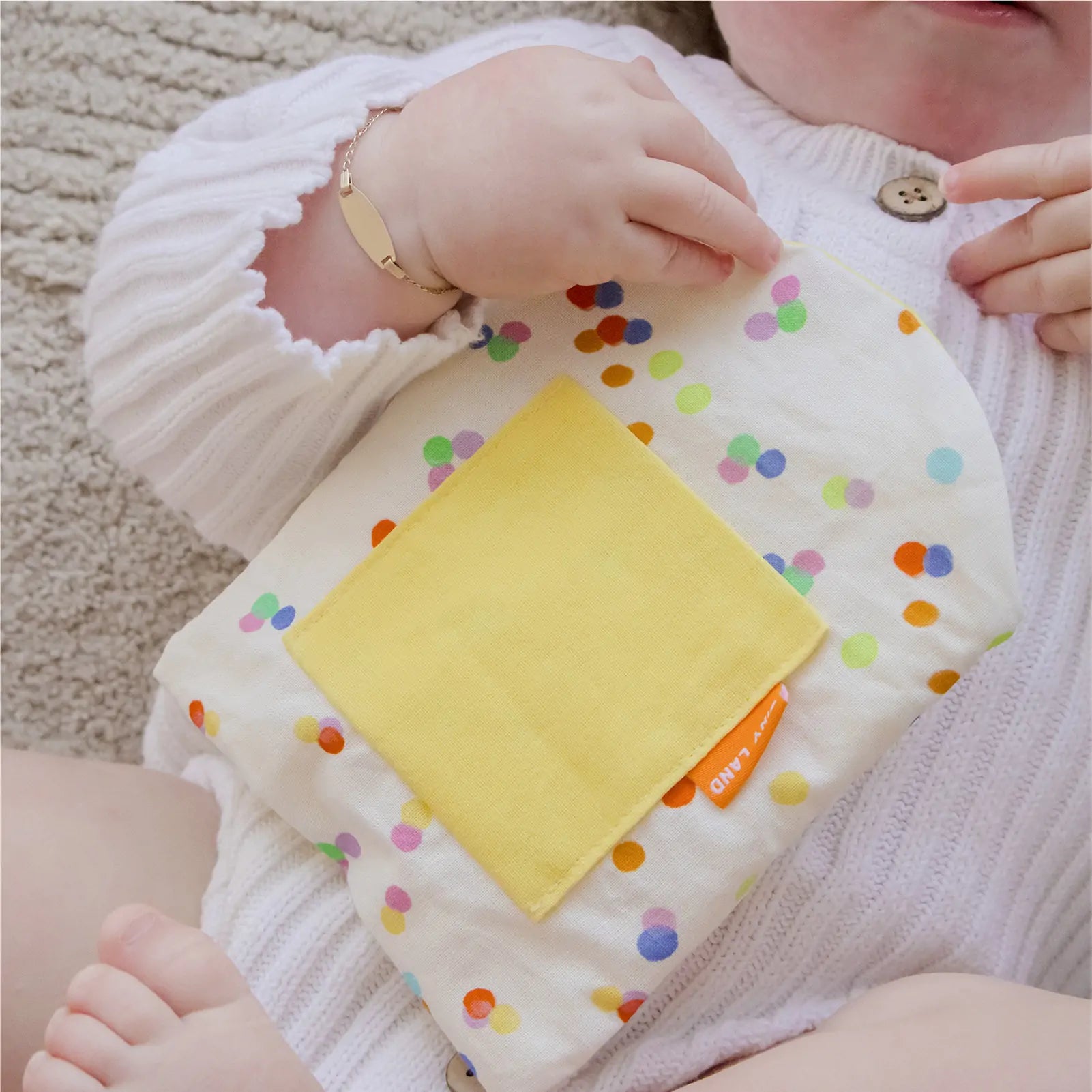A baby in a white knit outfit holds the Tiny Land® Montessori Toys for Infants (10–12 Months) pouch—white with colorful polka dots and a yellow pocket—a charming addition to any playset from Tiny Land, ideal for baby development.