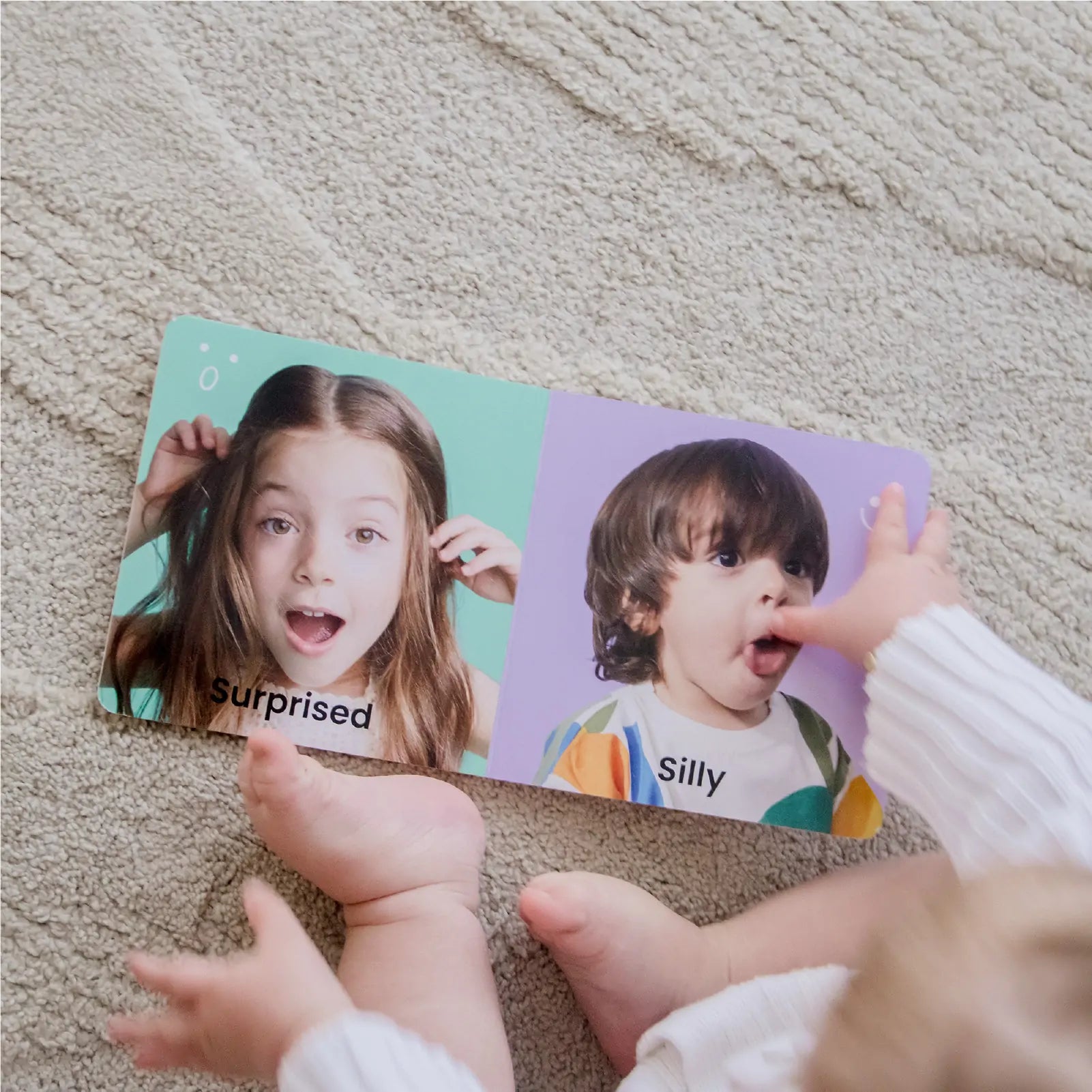 A baby sits on a carpet, exploring a Surprised and Silly board book page, surrounded by Tiny Land® Montessori Toys for Infants (10–12 Months) from Tiny Land that spark curiosity and support early learning.