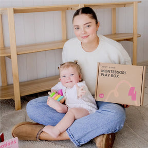 A woman sits on the floor holding a baby, who plays with colorful stacking rings from the Tiny Land® Montessori Toys for Infants (10–12 Months) set by Tiny Land, in their cozy, light-colored room.