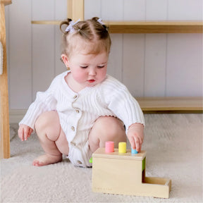 A baby with light brown hair and white bows, dressed in a white sweater and romper, sits on a carpet and plays with the Tiny Land® Montessori Toys for Infants (10–12 Months) by Tiny Land, featuring colorful wooden pegs.