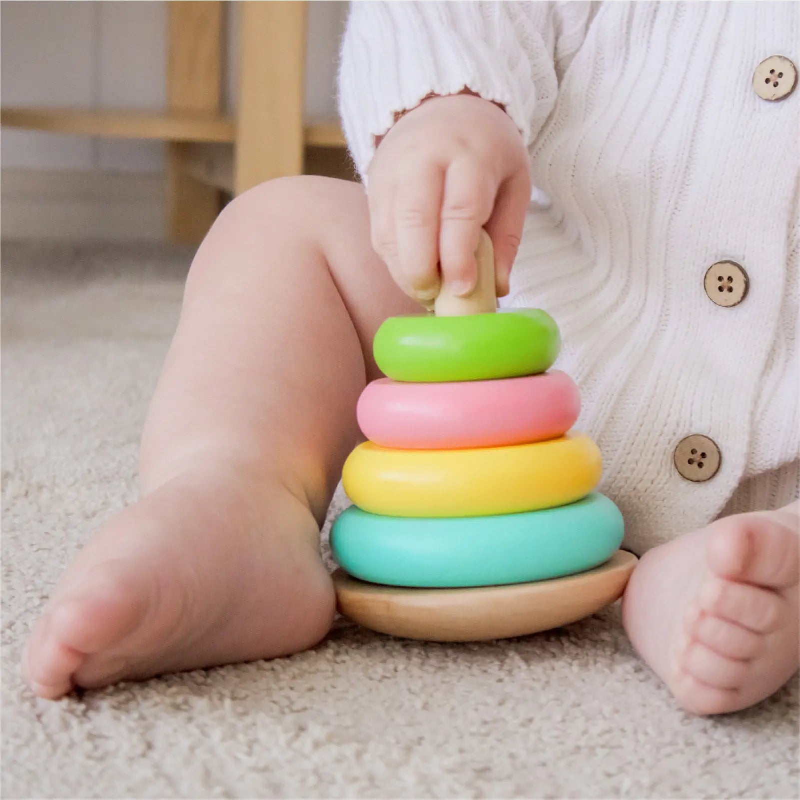 A baby in a white knit outfit sits on a carpet, stacking colorful rings from the Tiny Land® Montessori Toys for Infants (10–12 Months) playset by Tiny Land, using one hand to place a green ring on top.