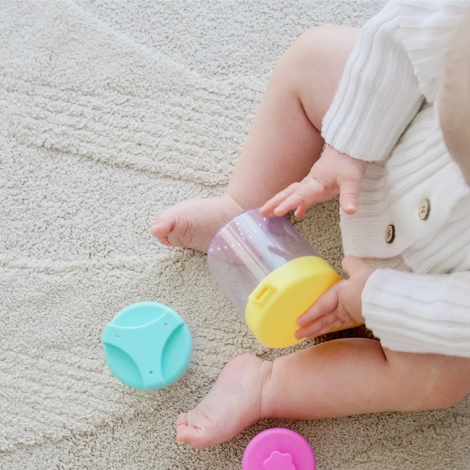 A baby in a white sweater sits on a light carpet, holding a clear snack container with a yellow lid. Nearby, Tiny Land® Montessori Toys for Infants (10–12 Months) support development and encourage exploration through colorful play.