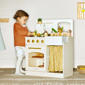 A young child stands beside the Tiny Land® Chef’s Corner Playset – Cream Playhouse + Cream Kitchen Set, pretending to cook with toy vegetables and accessories. The set features gold-colored knobs and a yellow patterned curtain in a softly lit room|white