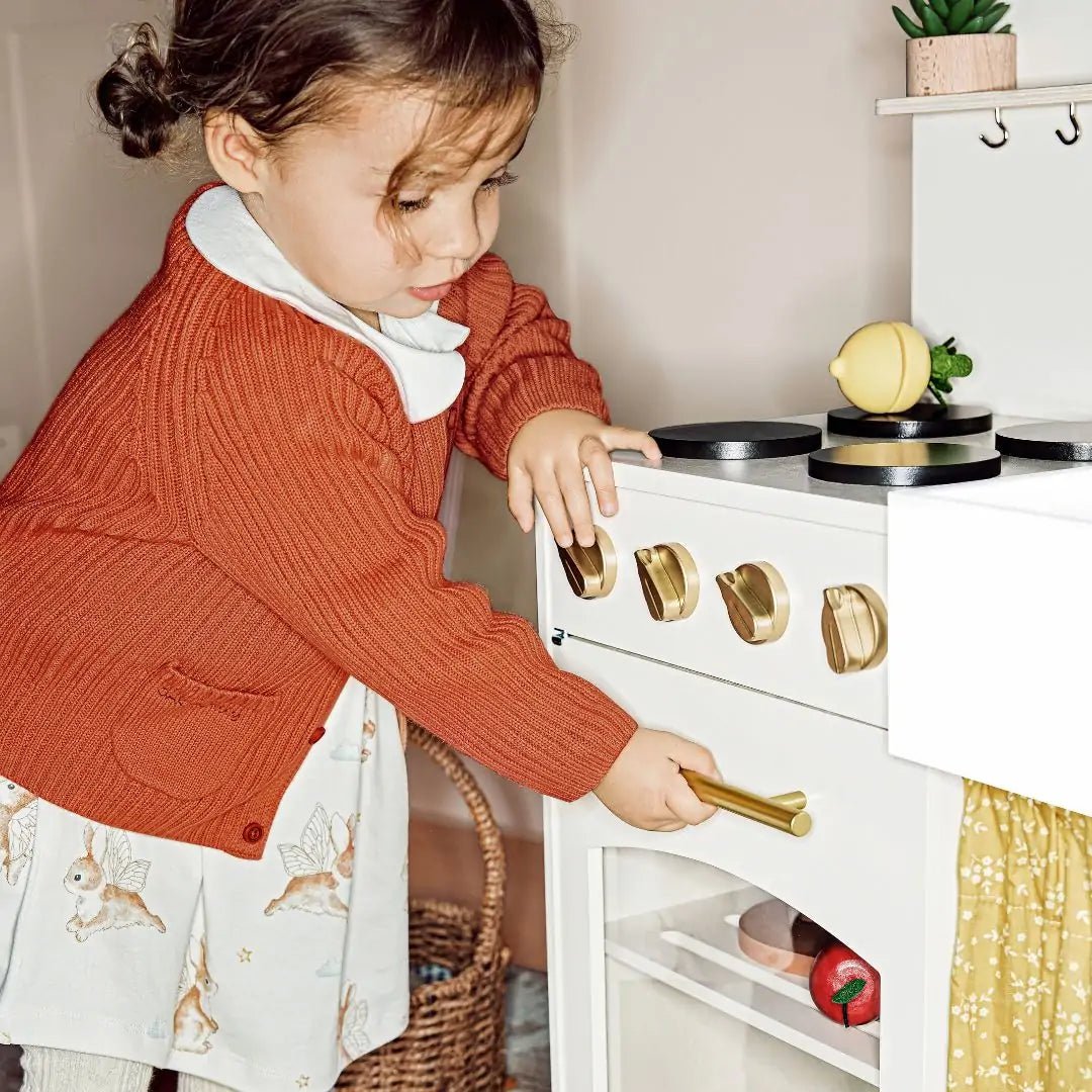 A young child in a rust-colored cardigan and printed dress enjoys imaginative play with the Tiny Land® Wooden Play Kitchen for Toddlers, turning the handle on the Honey Kitchenettes white oven while a toy lemon rests on the stove|white