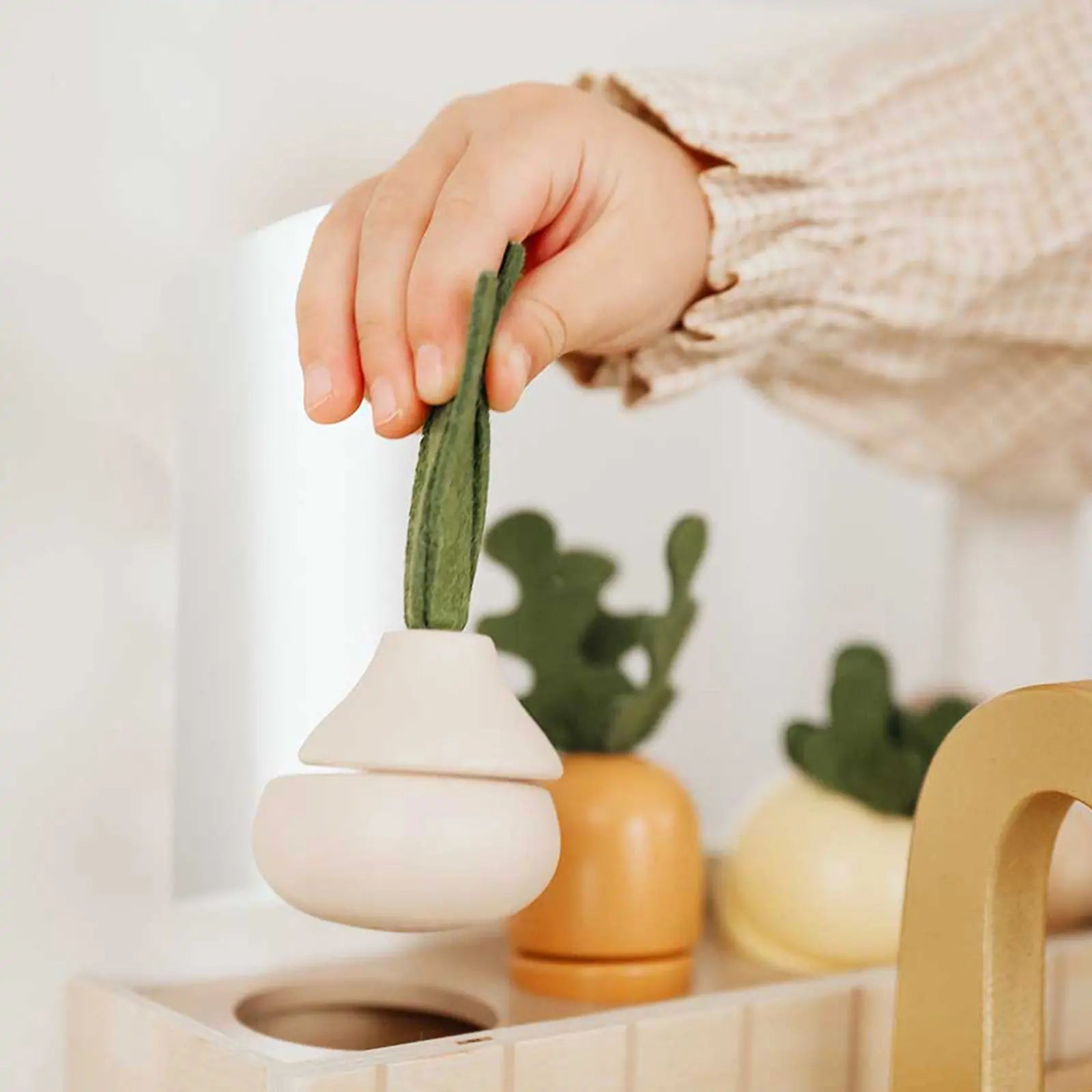 A child’s hand holds a plush vegetable toy—ideal for imaginative play with the Tiny Land® Wooden Play Kitchen for Toddlers. Other plush vegetables are seen on a light wooden surface near the Tiny Land Honey Kitchenette Pretend Cooking Set|white