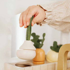 A child’s hand holds a plush vegetable toy—ideal for imaginative play with the Tiny Land® Wooden Play Kitchen for Toddlers. Other plush vegetables are seen on a light wooden surface near the Tiny Land Honey Kitchenette Pretend Cooking Set|white