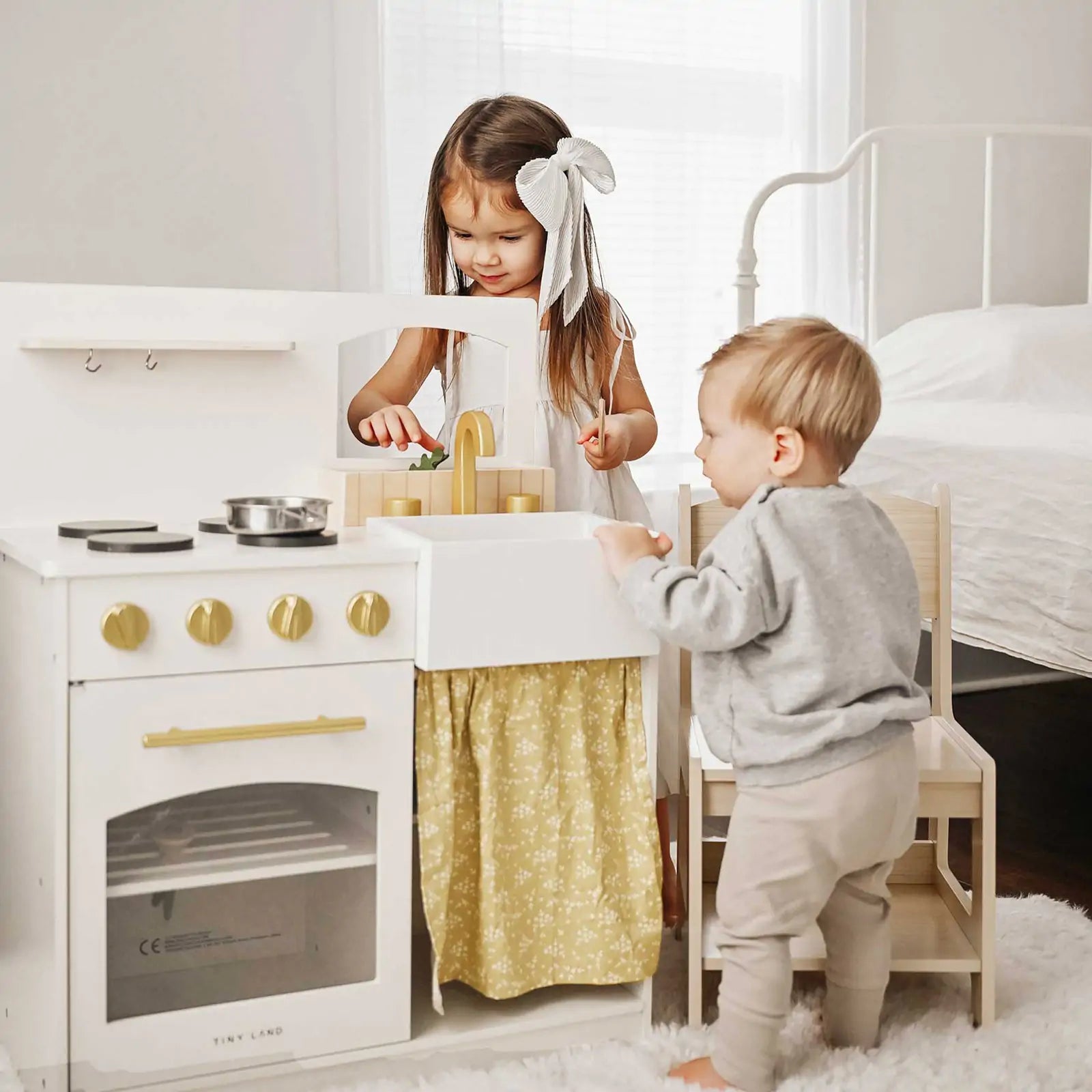 A young girl and toddler play with the Tiny Land® Chef’s Corner Playset – Cream Playhouse + Cream Kitchen Set in a bright bedroom, pretending to cook together as they enjoy their favorite kitchen set from Tiny Land|white