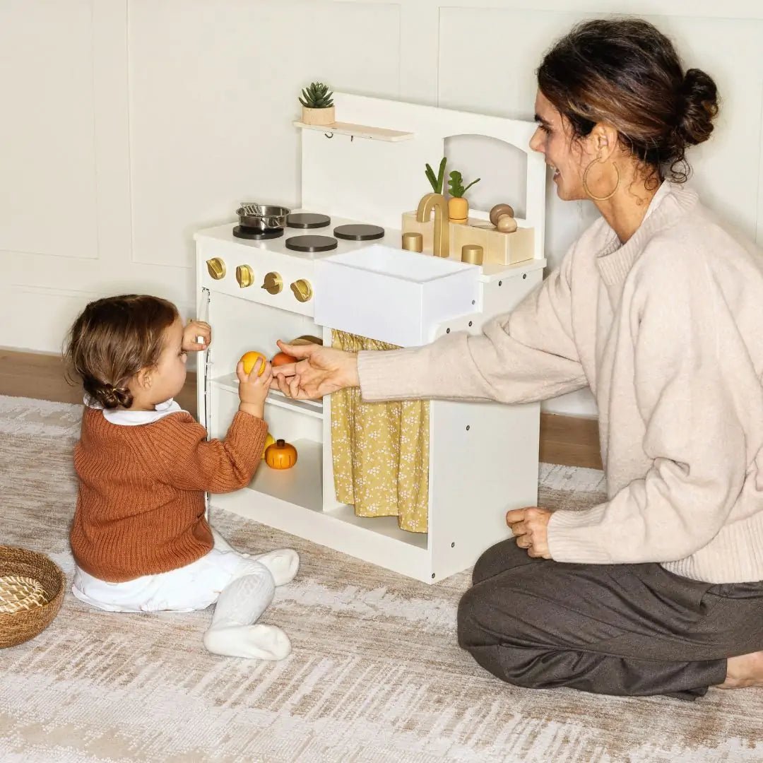 A woman kneels beside a toddler playing with the Tiny Land® Wooden Play Kitchen for Toddlers – Honey Kitchenette Pretend Cooking Set, both smiling as they enjoy imaginative pretend cooking and the child holds a toy orange|white