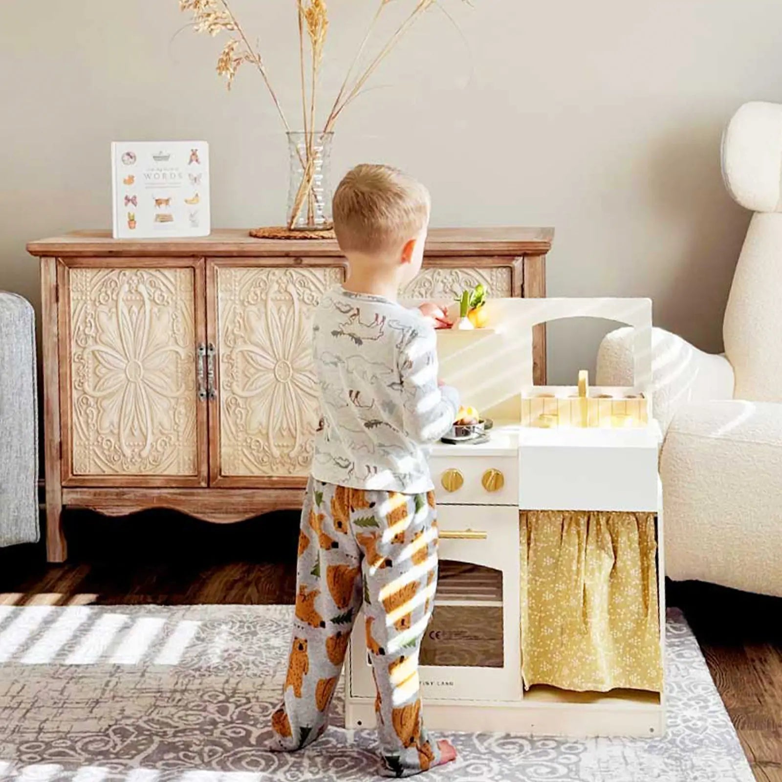 A young child in pajamas enjoys imaginative play with the Tiny Land® Wooden Play Kitchen for Toddlers – Honey Kitchenette Pretend Cooking Set, set in a sunlit living room with patterned carpet and a carved wooden cabinet behind|white