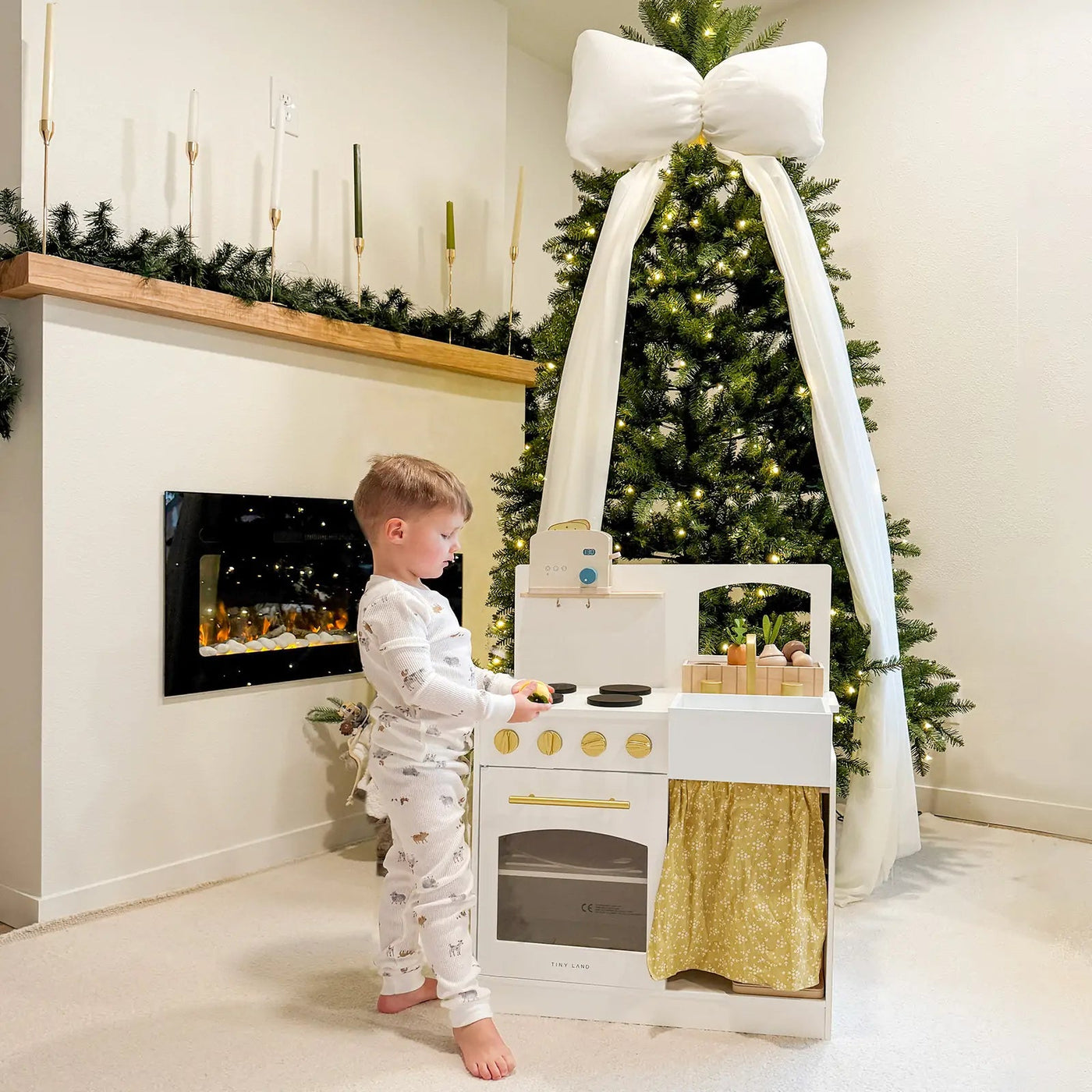 A young child in pajamas enjoys imaginative play with the Tiny Land® Wooden Play Kitchen for Toddlers – Honey Kitchenette Pretend Cooking Set, surrounded by Christmas decor including a lit tree with a white bow and a mantle adorned with greenery and candles|white