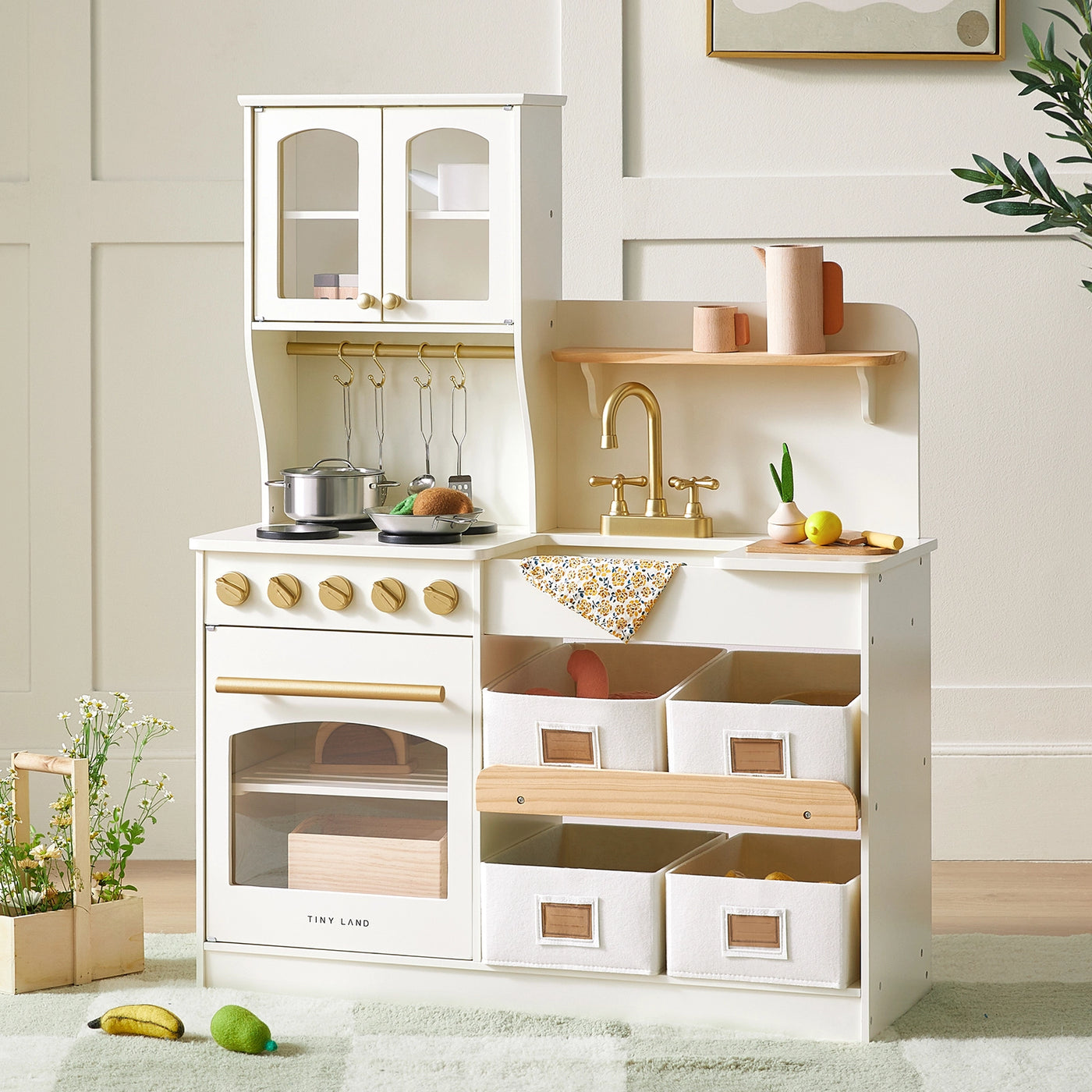 The Tiny Land® Wooden Play Kitchen with Storage by Tiny Land features a stove, oven, sink with golden faucet, hanging utensils, and six white bins. Pretend cookware and toy food are arranged on the counter and floor in a bright, cozy room|cream