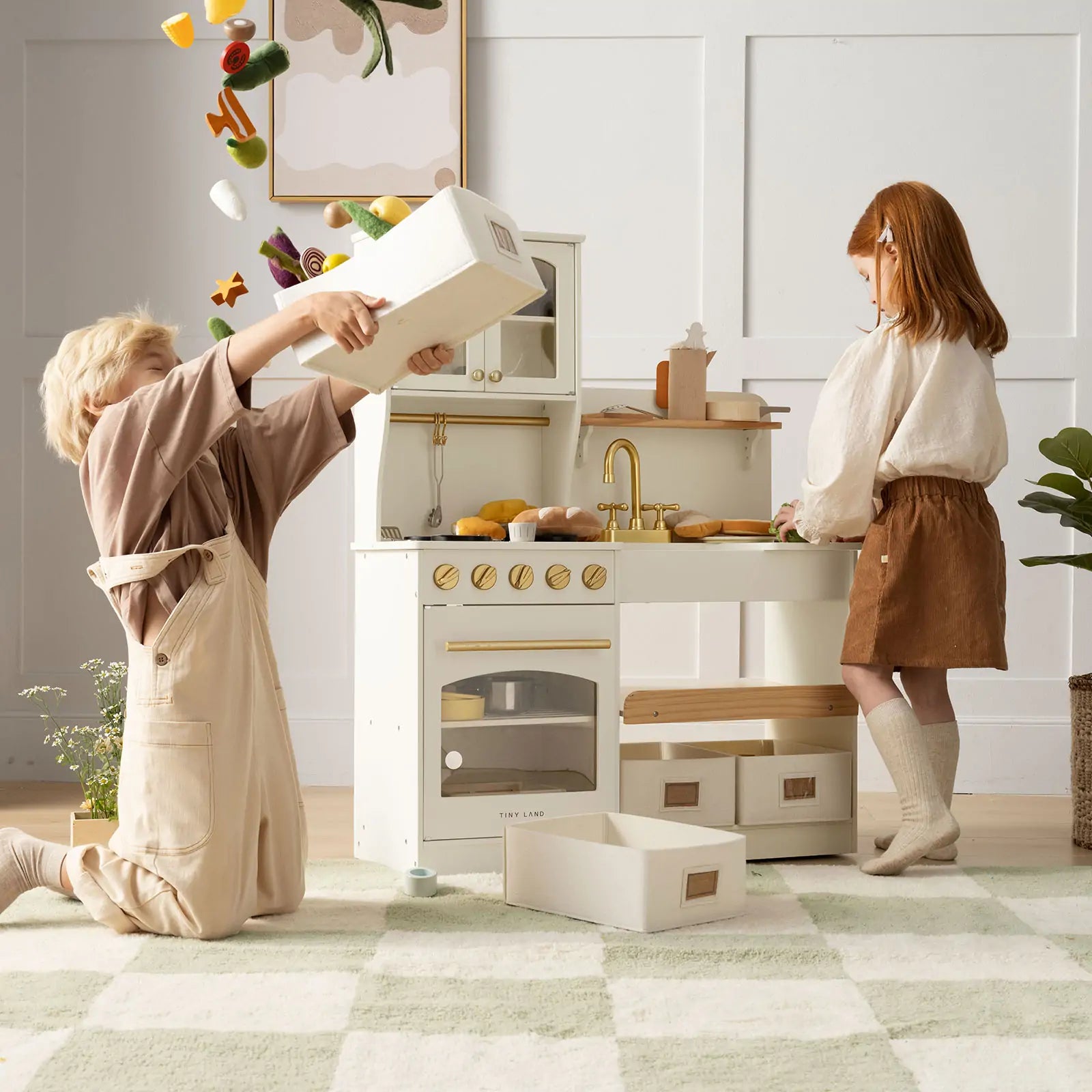 Two children enjoy imaginative play with the Tiny Land® Smart Storage Kitchen Cafe Set by Tiny Land; one kneels to dump out toy food while the other watches, surrounded by a wooden play kitchen and scattered pretend groceries|cream