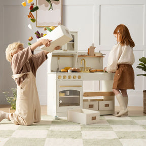 Two children enjoy imaginative play with the Tiny Land® Smart Storage Kitchen Cafe Set by Tiny Land; one kneels to dump out toy food while the other watches, surrounded by a wooden play kitchen and scattered pretend groceries|cream