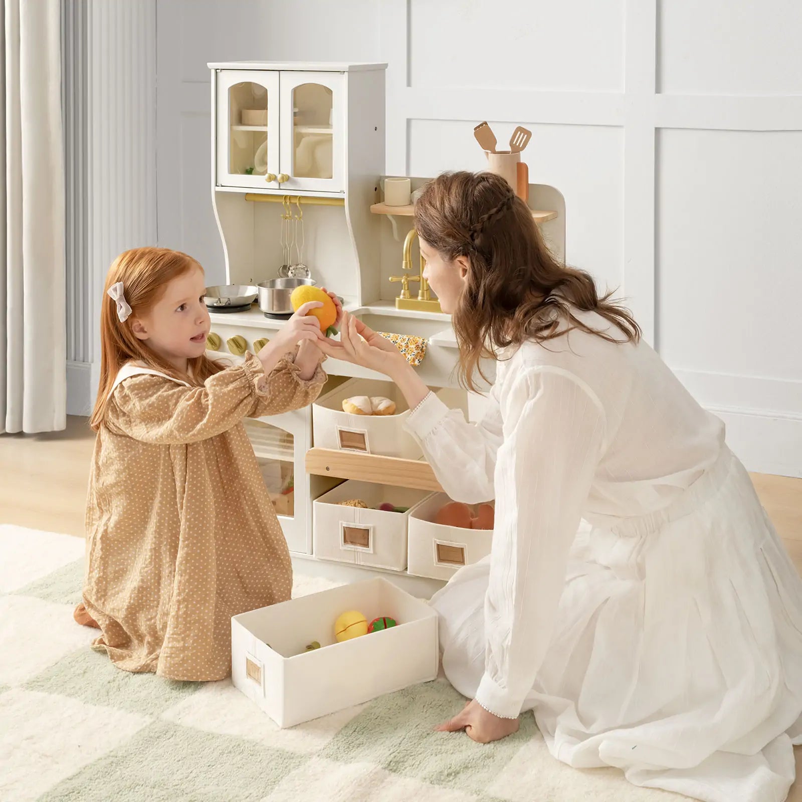 A young girl and a woman enjoy kitchen role-play on a light rug, using the Tiny Land® Wooden Play Kitchen with Storage for Creative Toddlers. The girl wears a brown dress and the woman a white one as they cook with pretend food and cookware|cream