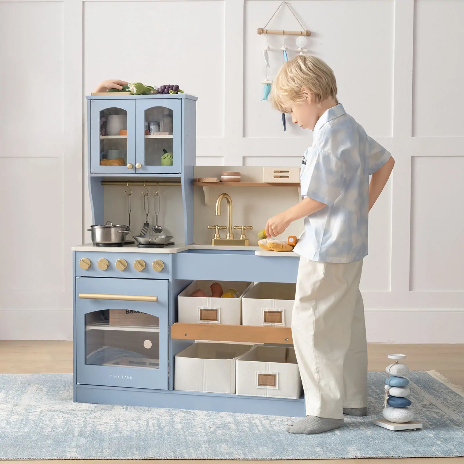 A young child enjoys imaginative play with the Tiny Land® Pretend Play Kitchen for Kids – Serenity Wooden Kitchen in blue & green, featuring a sink, stove, shelves, cabinets, and storage bins by Tiny Land in a tidy, well-lit room.