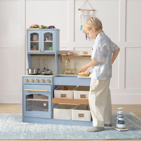A young child enjoys imaginative play with the Tiny Land® Pretend Play Kitchen for Kids – Serenity Wooden Kitchen in blue & green, featuring a sink, stove, shelves, cabinets, and storage bins by Tiny Land in a tidy, well-lit room.