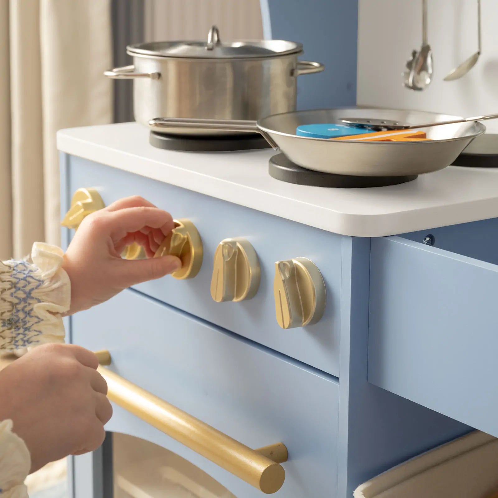 A child enjoys imaginative play with the Tiny Land® Pretend Play Kitchen for Kids – Serenity Wooden Kitchen in blue & green, turning gold knobs and using pretend metal pots and pans on the burners.