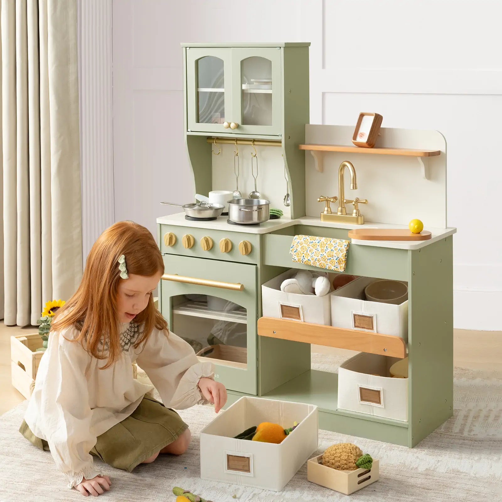 A young girl with red hair enjoys imaginative play beside the Tiny Land® Pretend Play Kitchen for Kids - Serenity Wooden Kitchen – Blue & Green, featuring pretend cookware, play food, utensils, a sink, and storage bins in natural light.