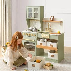 A young girl with red hair enjoys imaginative play beside the Tiny Land® Pretend Play Kitchen for Kids - Serenity Wooden Kitchen – Blue & Green, featuring pretend cookware, play food, utensils, a sink, and storage bins in natural light.