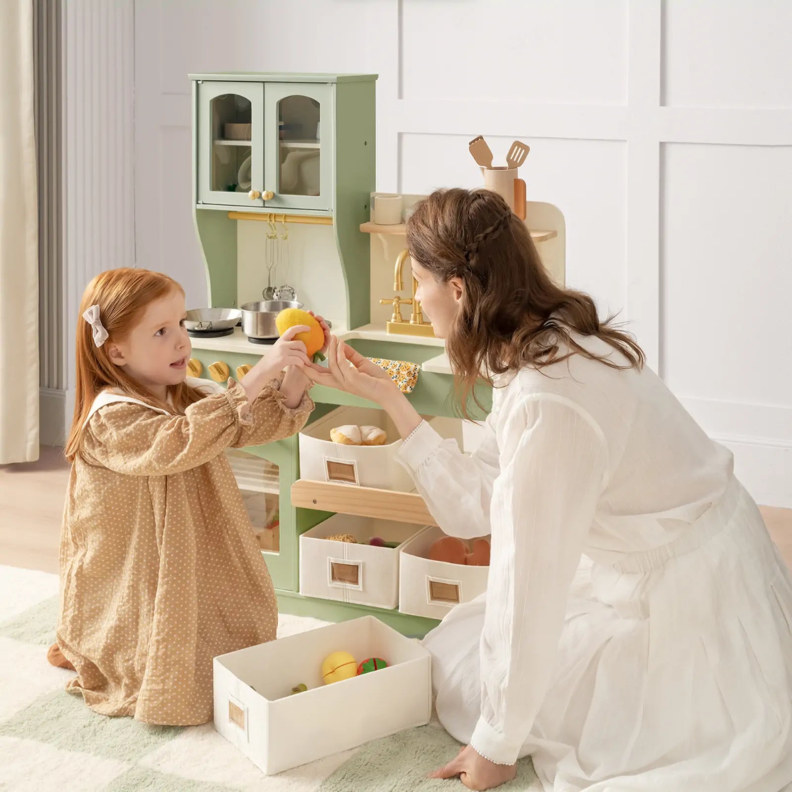 A woman and a young girl enjoy pretend play with the Tiny Land Little Green World Gift Set: Sage Dollhouse & Sage Storage Kitchen, surrounded by the bright, cheerful sage green play kitchen as they share toy fruit|sage