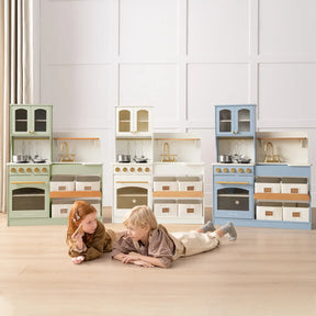 Two kids smile at each other on a light wood floor, playing with the Tiny Land Little Green World Gift Set: Sage Dollhouse & Sage Storage Kitchen—stylish, colorful sets with shelves, cabinets, and play accessories for endless pretend fun