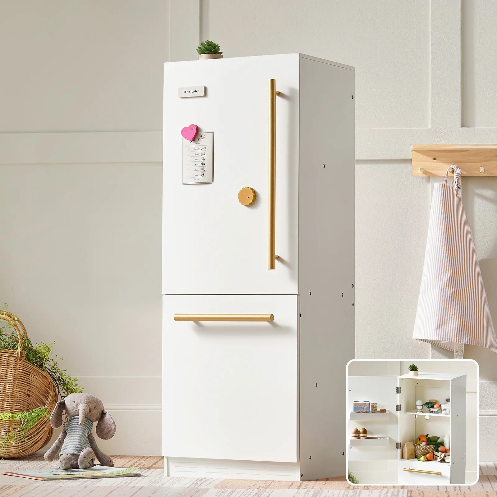 The Tiny Land® PopJoy Kitchen & Fridge Set features a white fridge with gold handles, a list held by a pink heart magnet, and shelves stocked with toy food. A stuffed elephant, basket, and apron complete this playful set by Tiny Land.
