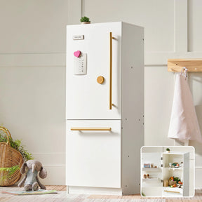The Tiny Land® PopJoy Kitchen & Fridge Set features a white fridge with gold handles, a list held by a pink heart magnet, and shelves stocked with toy food. A stuffed elephant, basket, and apron complete this playful set by Tiny Land.