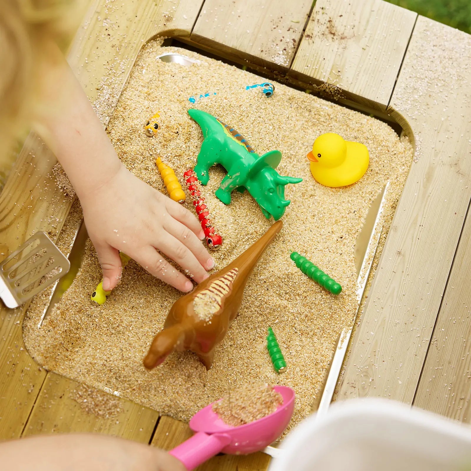 A child’s hand explores sensory play with toy dinosaurs, a yellow duck, colorful worms, and a pink scoop—perfect to use with the Tiny Land® Mud Kitchen - DuoPlay Outdoor Play Kitchen for Kids by Tiny Land.