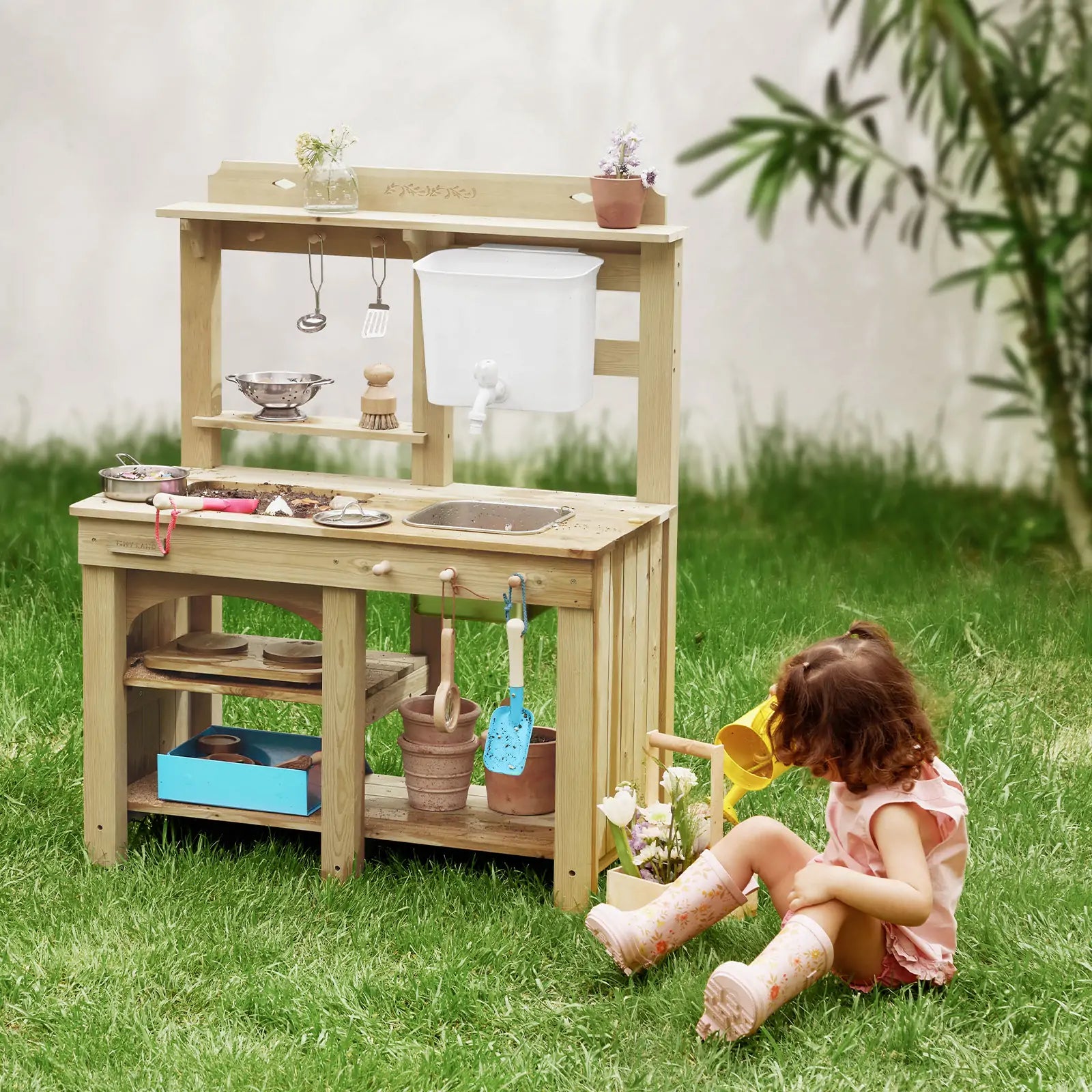 A young girl in pink boots enjoys hours of sensory play on green grass beside the Tiny Land® Mud Kitchen - DuoPlay Outdoor Play Kitchen for Kids, featuring utensils, a sink, and potted plants amidst lush garden greenery.